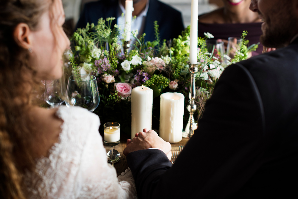 Couple holding hands during dinner at indoor wedding reception venues decorated with candles and flowers.